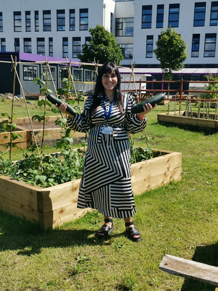 Julia Mottershead, a woman in a smart black and white striped dress, stands outside holding vegetables.