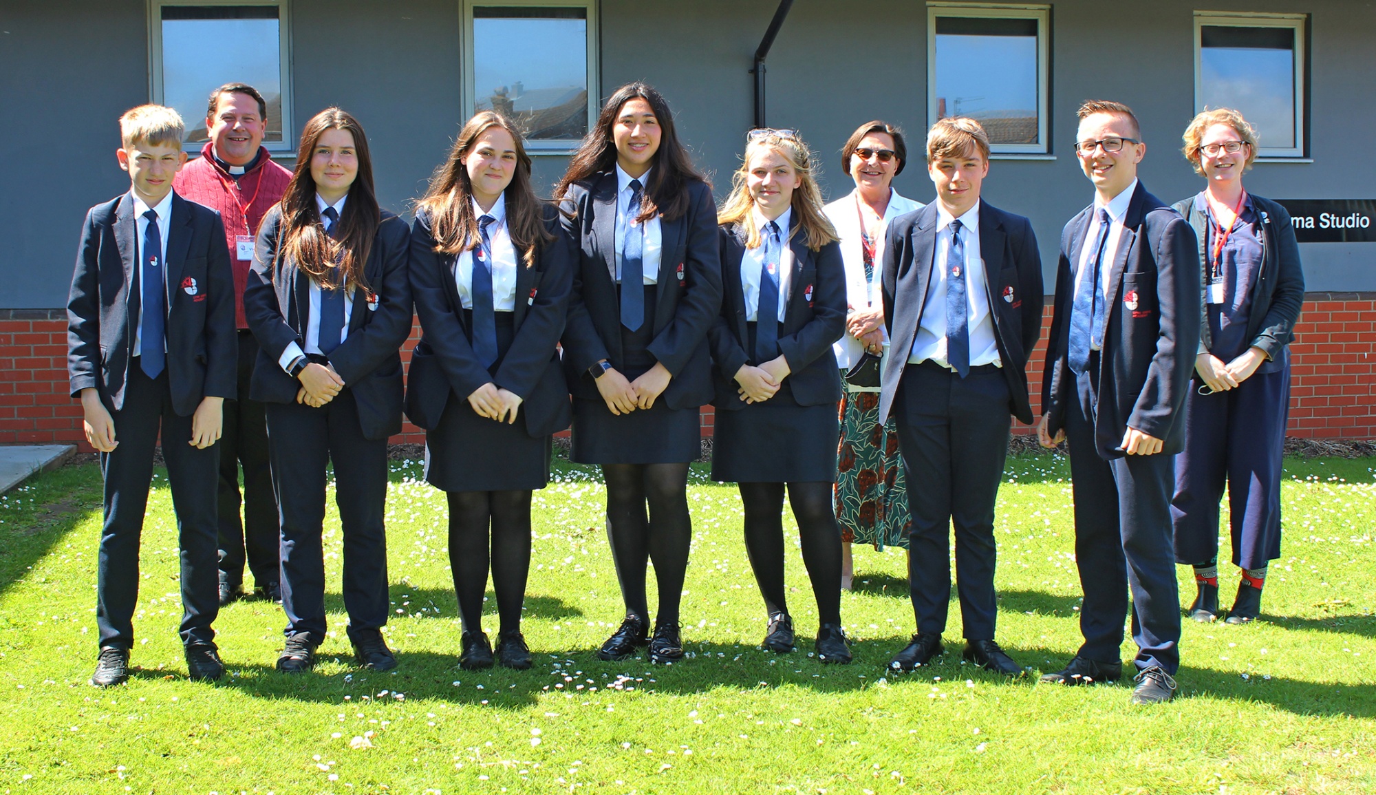 A group of pupils stand in two rows. outdoors, in front of a school building. There are three adults, the competition judges,, in the back row.