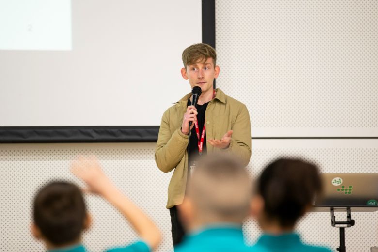 A speaker - Nade Ward - speaks into a microphone, in front of a group of young pupils