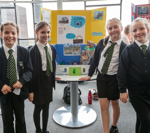 A group of young students in school uniform stand smiling in front of a handmade model of the coast.